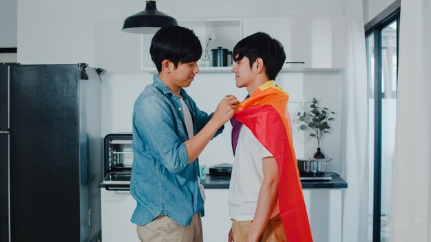 Asiatische homosexuelle Paare, die zu Hause Raum stehen und umarmen. Die jungen hübschen LGBTQ + Männer, die glücklich küssen, entspannen sich Rest verbringen zusammen romantische Zeit in der modernen Küche mit Regenbogenflagge am Haus morgens.
