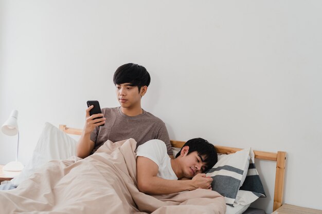 Asiatische homosexuelle Paare unter Verwendung des Handys zu Hause. Junge Asia LGBTQ + Mann glücklich entspannen Erholung nach dem Aufwachen, überprüfen Social Media, während sein Freund schlafen morgens auf dem Bett im Schlafzimmer zu Hause liegen.