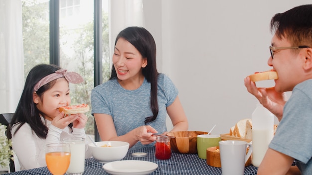 Asiatische japanische Familie frühstückt zu Hause. Die asiatische glückliche Mutter, die Erdbeermarmelade auf Brot für Tochter macht, essen Corn Flakes Getreide und Milch in der Schüssel auf Tabelle in der Küche morgens.