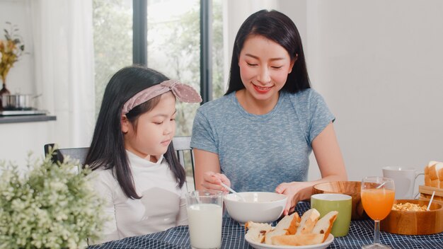 Asiatische japanische Familie frühstückt zu Hause. Glückliche Unterhaltung der asiatischen Mutter und der Tochter beim Essen des Brotes, des Orangensaftes, des Corn Flakesgetreides und der Milch auf Tabelle in der modernen Küche am Morgen.