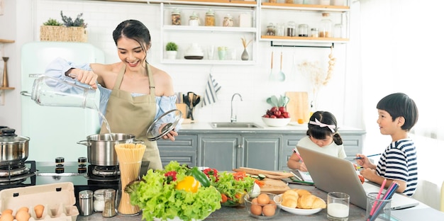 Asiatische junge alleinerziehende Mutter, die Essen zubereitet, während sie sich um ihr Kind in der Küche kümmert