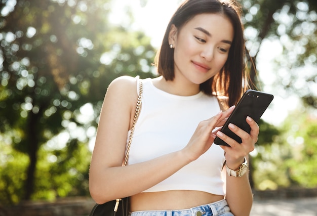 Asiatische junge Frau, die im Park steht und Nachricht auf Handy liest