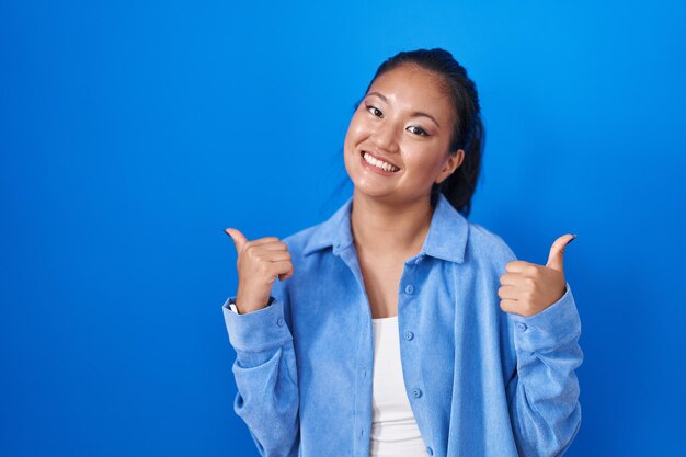 Asiatische junge Frau, die über blauem Hintergrund steht, Erfolgszeichen macht, positive Geste mit der Hand macht, Daumen hoch, lächelnd und glücklich. fröhlicher Gesichtsausdruck und Siegergeste.
