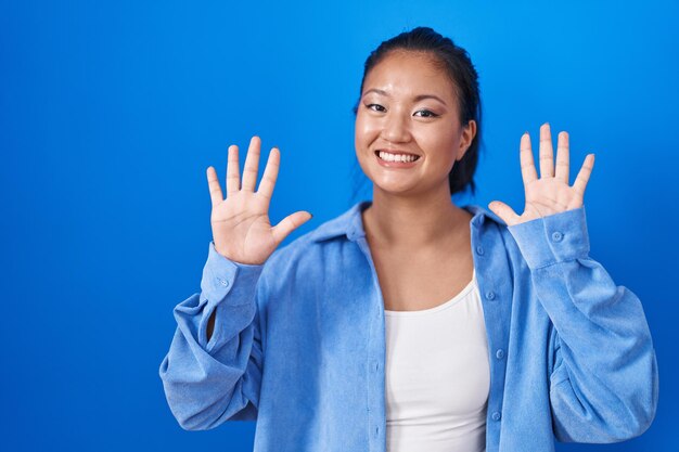 Asiatische junge Frau, die über blauem Hintergrund steht und mit den Fingern Nummer zehn nach oben zeigt, während sie selbstbewusst und glücklich lächelt.