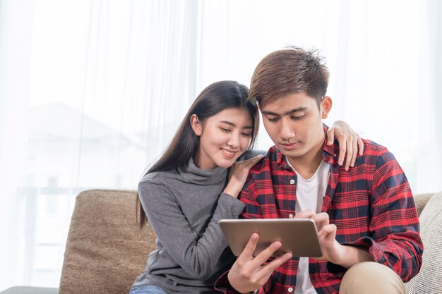 Asiatische junge Frau und gutaussehender Mann, die auf Couch unter Verwendung des Geräts sitzt