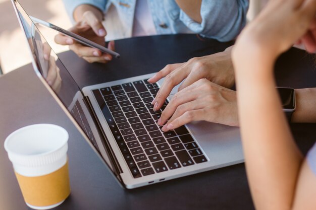 Asiatische junge Frauen, die an Laptop unter Verwendung und Schauen des Smartphone und trinkender Kaffee arbeiten