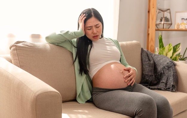 Asiatische junge schwangere Frau, die Bauchschmerzen hat, sitzt zu Hause auf einer Couch im Wohnzimmer