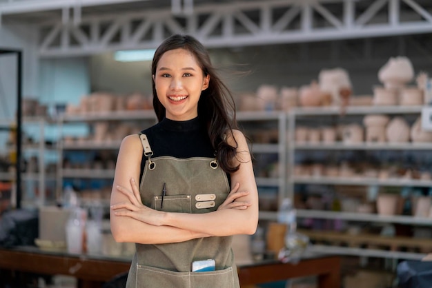 Asiatische Keramikerin mit Schürze, selbstbewusster Brust in ihrer Werkstatt, Tonskulpturenstudio mit positivem, lächelndem, herzlichem Willkommen, bereit, eine neue Fabrikkeramikwerkstatt mit einer neuen Sammlung von Arbeiten zu starten