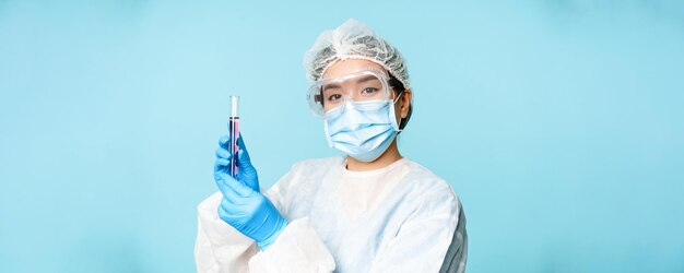 Asiatische Krankenschwester oder Laborantin in persönlicher Schutzausrüstung, die ein Testprobenröhrchen zeigt, das in einer medizinischen Gesichtsmaske auf blauem Hintergrund steht