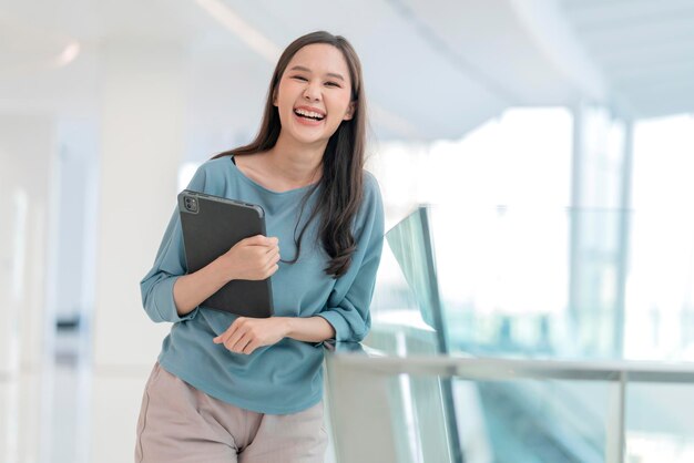 Asiatische lächelnde fröhliche weibliche digitale Nomaden-Handgriff-Tablet-Gerät Blick in die Kamera Porträt shothappiness lächelnde asiatische Frau, die im Büro-College-Korridor mit positiver lächelnder Haltung steht