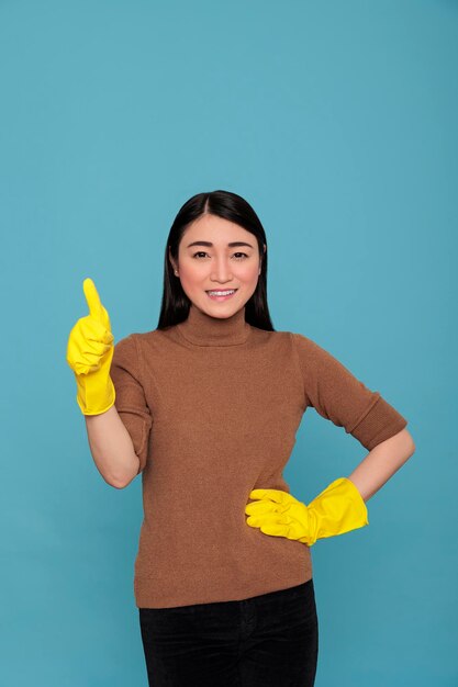 Asiatische lächelnde, glückliche asiatische Hausmädchen mit dem Daumen nach oben und mit gelben Handschuhen für die Sicherheit der Hände, Reinigungskonzept für zu Hause, zufriedene lachende und positive Geisteshaltung Frau, die bereit ist, die täglichen Aufgaben zu erledigen