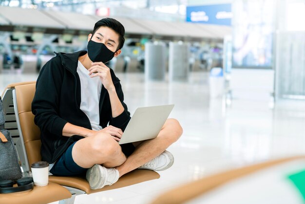 Asiatische männliche lässige tuch tragen virenschützende gesichtsmaske sitzen mit sozialem sitz, der den neuen normalen lebensstil distanziert, genießen handarbeit verwenden laptop im sicherheitsreisekonzept des flughafenterminals