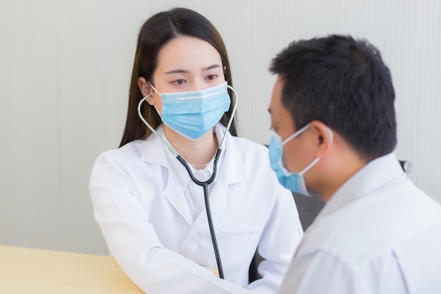 Asiatische männliche patienten werden auf seinen gesundheitszustand untersucht, während die ärztin ein stethoskop verwendet, um die herzfrequenz zu hören