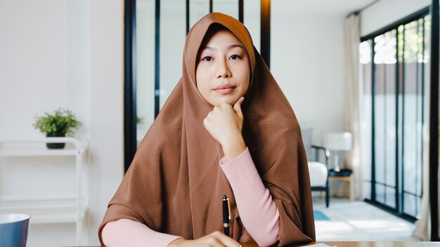 Asiatische muslimische Dame trägt Hijab mit Computer-Laptop. Sprechen Sie mit Kollegen über den Plan in Videoanrufen, während Sie von zu Hause aus im Wohnzimmer arbeiten.