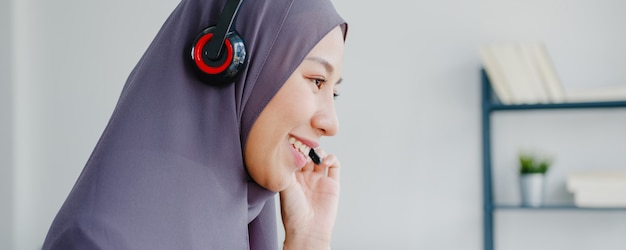 Asiatische muslimische Dame trägt Kopfhörer-Uhr-Webinar Hören Sie den Online-Kurs per Konferenz-Videoanruf zu Hause.