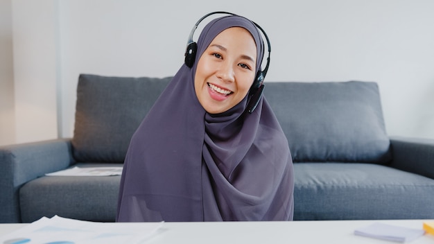 Kostenloses Foto asiatische muslimische dame tragen kopfhörer mit computer-laptop sprechen mit kollegen über den plan in videoanrufbesprechungen, während sie von zu hause aus im wohnzimmer arbeiten