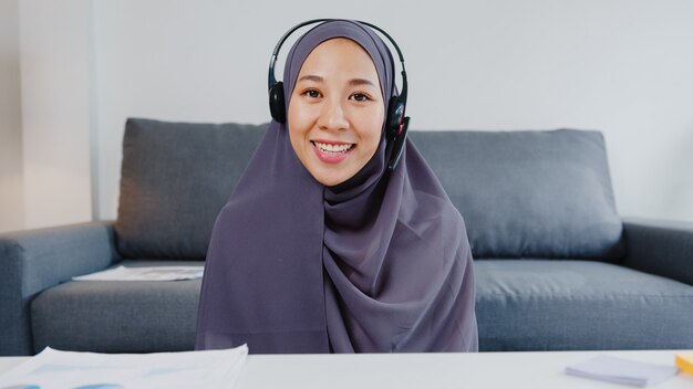 Asiatische muslimische dame tragen kopfhörer mit computer-laptop sprechen mit kollegen über den plan in videoanrufbesprechungen, während sie von zu hause aus im wohnzimmer arbeiten