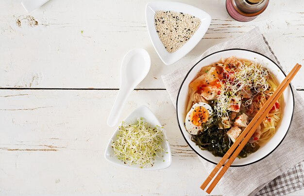 Asiatische Nudeln Miso Ramen mit Kohl Kimchi, Meerespflanze, Ei, Pilzen und Käsetofu in der Schüssel auf weißem Holztisch.