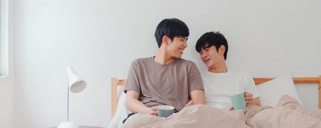 Asiatische Paare der homosexuellen Männer, die die schöne Zeit am modernen Haus haben sprechen. Das junge glückliche Asien-Liebhaber LGBTQ + Mann entspannen sich Restgetränkkaffee, nachdem er morgens beim Lügen auf Bett im Schlafzimmer am Haus aufgewacht ist.