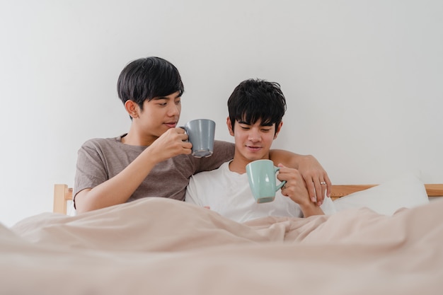 Asiatische Paare der homosexuellen Männer, die die schöne Zeit am modernen Haus haben sprechen. Das junge glückliche Asien-Liebhabermann entspannen sich Restgetränkkaffee nach wachen beim morgens liegen auf Bett im Schlafzimmer am Haus auf.