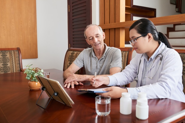 Asiatische Ärztin, die mit älterem kaukasischem Patienten während des Hausbesuchs spricht