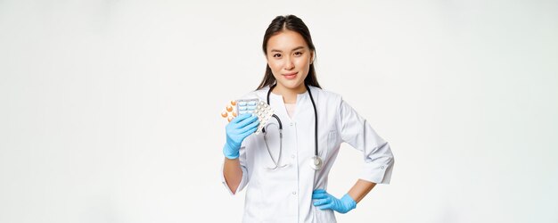 Asiatische Ärztin, die Pillenmedikamente zeigt, die Vitamine halten und Gummihandschuhe tragen, die über weißem Hintergrund stehen