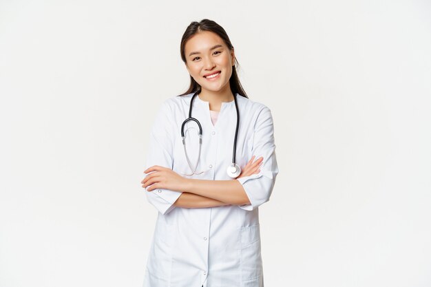 Asiatische Ärztin, Ärztin in medizinischer Uniform mit Stethoskop, Kreuzarme auf der Brust, lächelnd und professionell aussehend, weißer Hintergrund