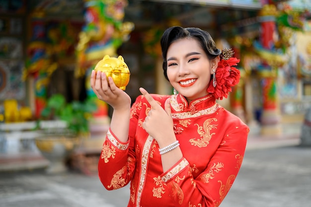 Asiatische schöne Frau des Porträts, die ein Cheongsam trägt, lächelt und posiert mit goldenem Sparschwein am Schrein am chinesischen Neujahrsfest