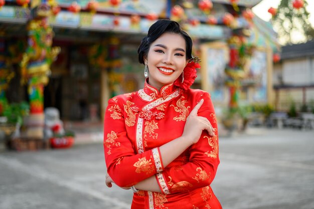 Asiatische schöne Frau des Porträts, die ein Cheongsam trägt, lächelt und posiert mit Gratulationsgeste am Schrein zum chinesischen Neujahr