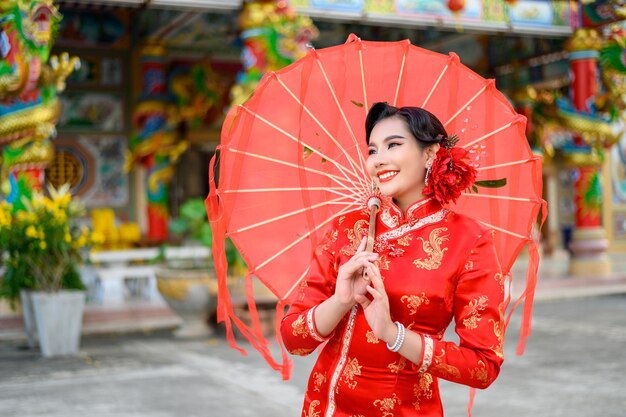 Asiatische schöne Frau des Porträts, die ein Cheongsam trägt, lächelt und posiert mit rotem Regenschirm aus Papier am Schrein am chinesischen Neujahrsfest