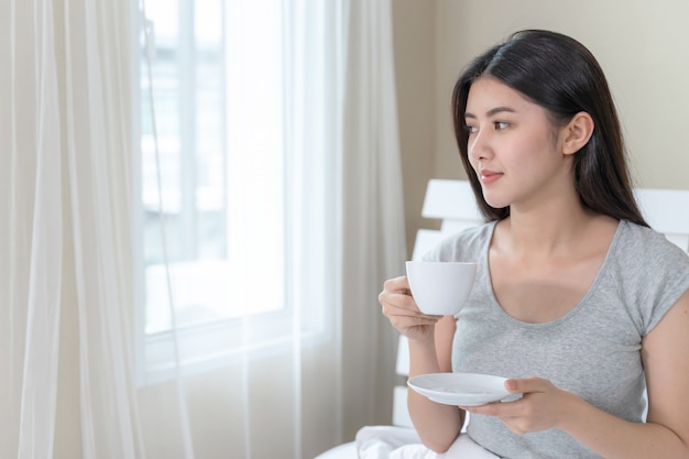 Asiatische Schönheit, die auf Bett im Schlafzimmer sitzt und in der Hand Kaffeetasse hält