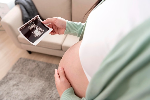 Asiatische schwangere Frau, die zu Hause ein Ultraschallbild auf dem Bauch hält Schwangerschaftsmutterschaftsvorbereitungs- und -erwartungskonzept