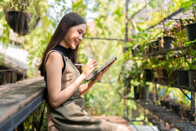 Asiatische weibliche Gärtnerin in Schürze, die in einem Garten im Freien zu Hause arbeitet. Gärtnerin mit Tablet-Computer zur Einstellung des Wassertropfensystems im Gewächshaus