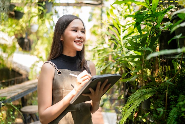 Asiatische weibliche Gärtnerin in Schürze, die in einem Garten im Freien zu Hause arbeitet. Gärtnerin mit Tablet-Computer zur Einstellung des Wassertropfensystems im Gewächshaus