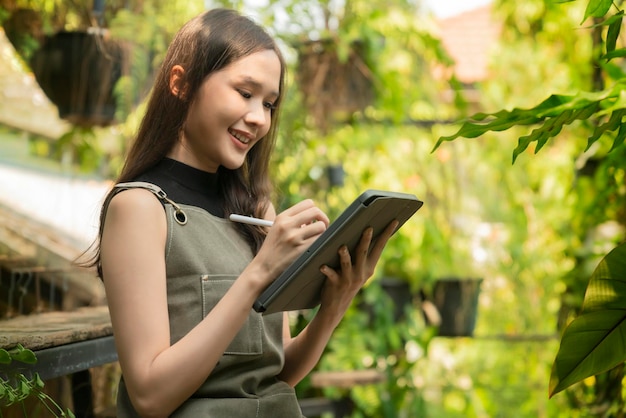 Asiatische weibliche Gärtnerin in Schürze, die in einem Garten im Freien zu Hause arbeitet. Gärtnerin mit Tablet-Computer zur Einstellung des Wassertropfensystems im Gewächshaus