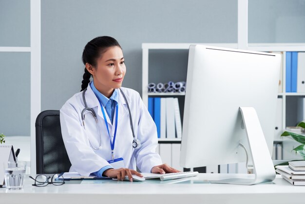 Asiatischer Doktor mit Stethoskop um den Hals, der im Büro sitzt und an Computer arbeitet