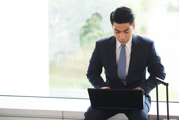 Asiatischer Geschäftsmann, der auf Fensterbrett sitzt, wenn der Koffergriff darstellt und an Laptop arbeitet