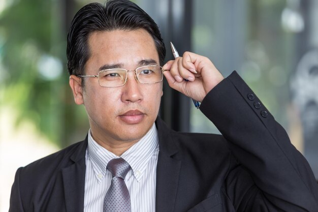Asiatischer Geschäftsmann im Anzug, der die Brille mit Stift berührt