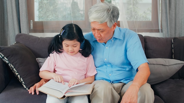 Asiatischer Großvater entspannen sich zu Hause. Der ältere Chinese, der glückliche Großvater entspannen sich mit jungem Enkelinmädchen genießen, Bücher zu lesen und Hausarbeit zusammen im Wohnzimmerkonzept zu erledigen.