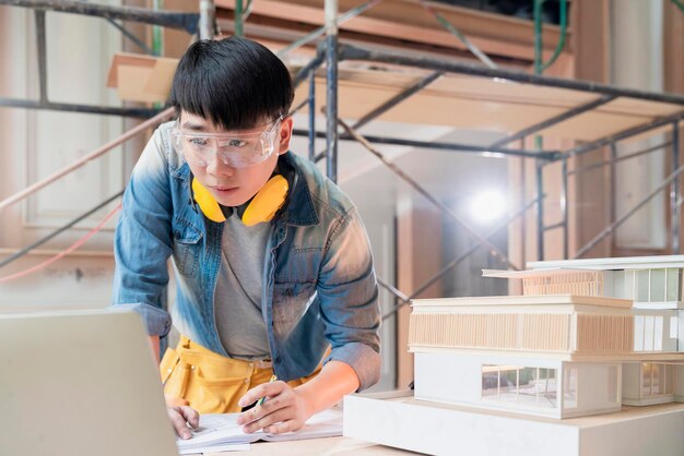 Asiatischer ingenieur männlich mit einheitlichem arbeiten mit laptop und hausmodellanalyse bauergebnis mit anwendungstechnologie am standort bauhaus renovierung und verbesserungshintergrund