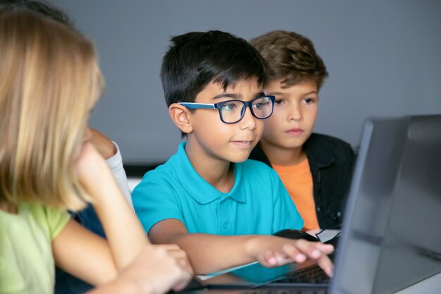 Asiatischer Junge, der auf Laptoptastatur und Klassenkameraden tippt, die am Tisch sitzen, ihn beobachten und Aufgabe zusammen erledigen