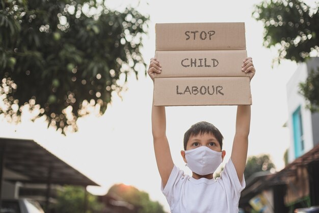 Asiatischer junge in medizinischer maske, der brieftafel anhebt, sagt kampagne „stopp kinderarbeit“