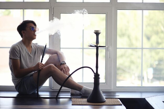 Asiatischer junger mann mit sonnenbrille raucht eine wasserpfeife am fenster