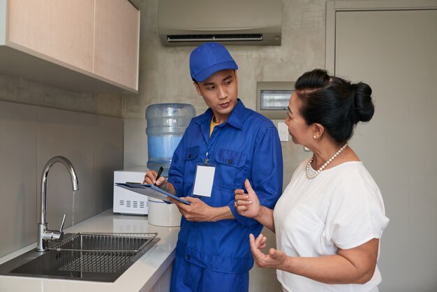 Asiatischer männlicher Klempner in der Uniform sprechend mit älterem weiblichem Hausbesitzer in der Küche