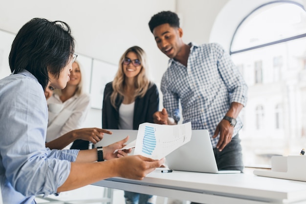 Asiatischer Manager in der Armbanduhr, die an seinem Arbeitsplatz sitzt und Papier mit Grafik hält. Innenporträt des afrikanischen Programmierers im karierten Hemd, das mit anderen Angestellten im Büro spricht.