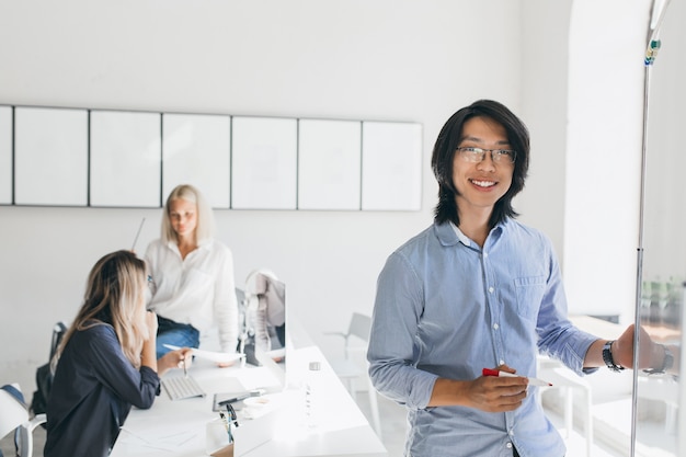 Asiatischer Manager mit aufrichtigem Lächeln, das neben Flipchart aufwirft, während Mädchen sprechen