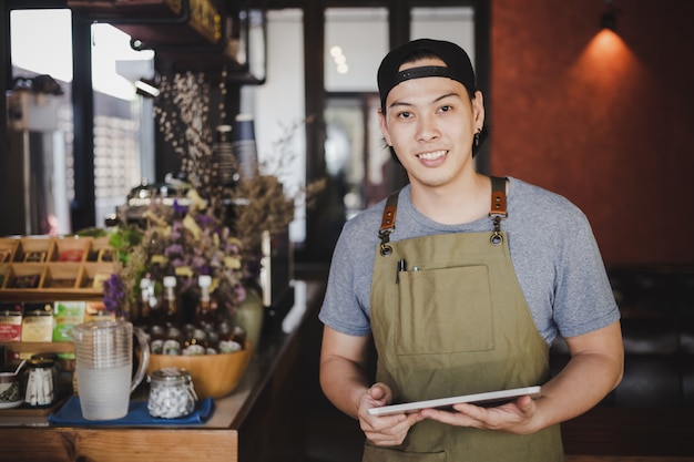 asiatischer Mann Barista, der Tablette für die Prüfung der Bestellung vom Kunden auf Kaffeecafé hält.