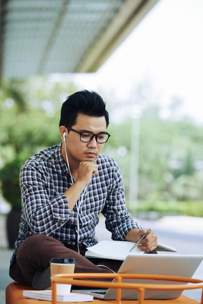 Asiatischer Mann, der draußen auf Bank mit Laptop sitzt und auf Online-Webinar hört