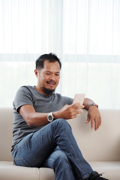 Asiatischer Mann, der gemächlich auf Couch mit den gekreuzten Beinen sitzt und Smartphone verwendet