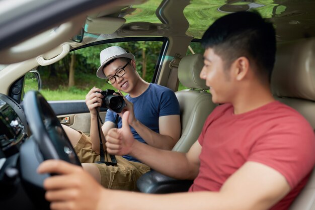 Asiatischer Mann, der im Auto hinter Lenkrad sitzt und für Freund mit Kamera aufwirft